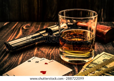 Glass of whiskey and playing cards with revolver and money on the wooden table. Angle view, image vignetting and the orange-blue toning, identification cards ace Russian letter