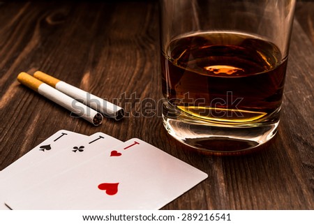 Glass of whiskey and playing cards with two cigarettes on the wooden table. Close up view