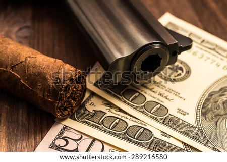 Barrel of a revolver with cuban cigar and money on the wooden table. Close up view, focus on the cuban cigar
