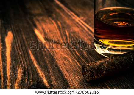 Glass of whiskey and cuban cigar on a wooden table. Close up view, focus on the cuban cigar, image vignetting and the orange-blue toning