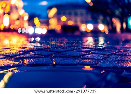 The bright lights of the evening city after rain, headlights of the cars riding straight. View from the pavement level next to the roadside puddle, image in the orange-blue toning