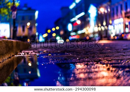 The bright lights of the evening city after rain, street with cars. View from the pavement level next to the roadside puddle, in blue tones
