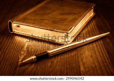 Golden pen with a leather diary on a mahogany table. Image vignetting and the yellow-orange toning