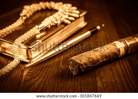 Golden pen with a leather diary and cuban cigar with jewellery on a mahogany table. Focus on the cuban cigar, image in the yellow-orange toning