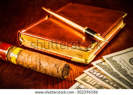 Money with a leather diary and cuban cigar with golden pen on a mahogany table. Focus on the cuban cigar, image vignetting and hard tones