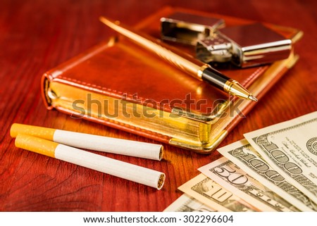 Money with a leather diary and cigarettes with golden pen on a mahogany table. Focus on the cigarettes