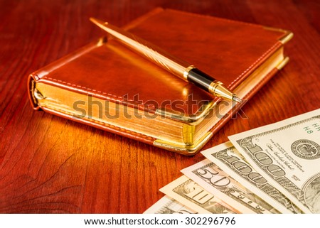 Golden pen on a leather diary with money on a mahogany table