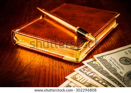 Golden pen on a leather diary with money on a mahogany table. Image vignetting and hard tones