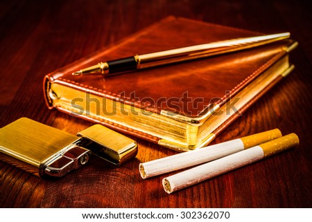 Leather diary and cigarettes with open golden lighter and pen on a mahogany table. Focus on the cigarettes, image vignetting and hard tones