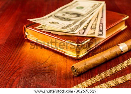 Money with a leather diary and cuban cigar with jewellery on a mahogany table. Focus on the cuban cigar