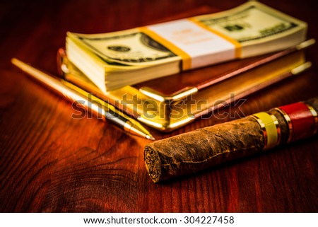 Pack of dollars with a leather diary and cuban cigar with golden pen on a mahogany table. Focus on the cuban cigar, image vignetting and hard tones