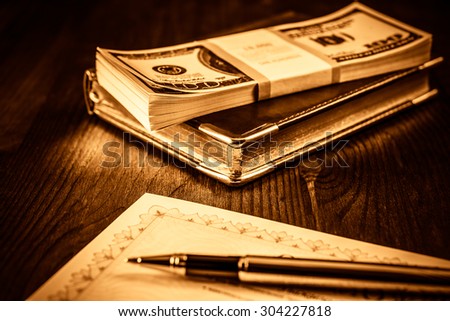Pack of dollars on the leather diary with bank check and golden pen on the table. Focus on the leather diary, image vignetting and yellow-orange toning
