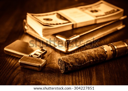 Pack of dollars and golden lighter with a leather diary and cuban cigar on a mahogany table. Shallow depth of field, focus on the cuban cigar, image vignetting and yellow-orange toning