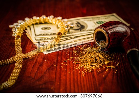 Tube for smoking tobacco and money with jewellery on a wooden table. Focus on the tube, image vignetting and hard tones