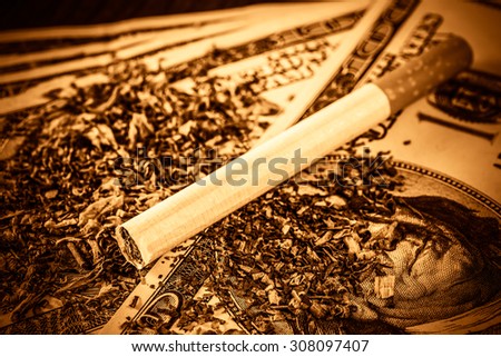 Cigarette and tobacco leaves lie scattered on the dollar bills on a wooden table. Close up view, image vignetting and hard yellow-orange toning