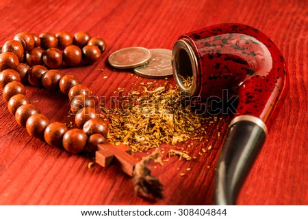 Tube for smoking tobacco with money and rosary with a cross on a wooden table. Close up view, focus on the tube