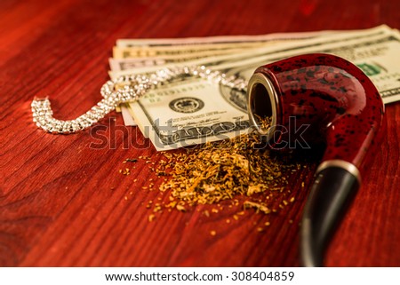 Tube for smoking tobacco and money with jewellery on a wooden table. Focus on the tube