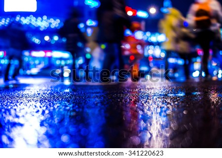 Rainy night in the big city, pedestrians cross the busy intersection in the light of shop windows. View from the level of asphalt, in blue tones