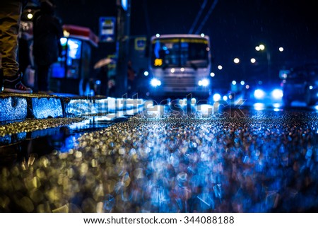 Rainy night in the big city, people standing at the bus stop waiting for the bus. View from the level of asphalt, in blue tones