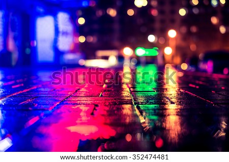 Rainy night in the big city, light from the night club and the windows of the house is reflected in the asphalt. View from the sidewalk level paved with bricks, image in the yellow-blue toning