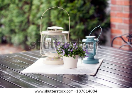 Moody ornamental arrangement with plant with blue flowers and lamps on a wooden table with metallic chairs and brick and green texture in the background