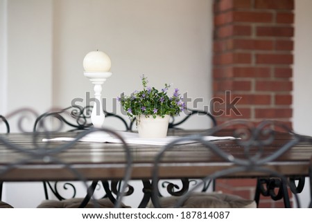 Moody ornamental arrangement with plant with blue flowers and candle on a wooden table with metallic chairs and brick texture in the background