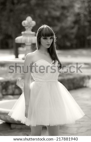 Black and white photo of a beautiful, caucasian, young, long haired woman posing in the park, wearing a white dress, looking happy