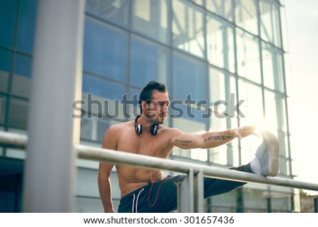 Young man listening to the music and stretching his leg after a heavy workout. Wearing tracksuits without shirt. Selective focus.