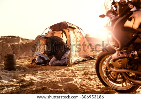Middle aged motorcyclist relaxing after a long ride on motorbike. Checking his travel map in tent. Lens flare. Warm colors. Selective focus.