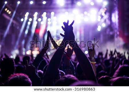 Audience with hands raised at a music festival and lights streaming down from above the stage. Soft focus, blurred movement.