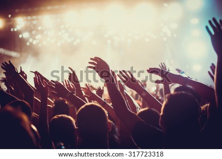 Audience with hands raised at a music festival and lights streaming down from above the stage. Soft focus, blurred movement.