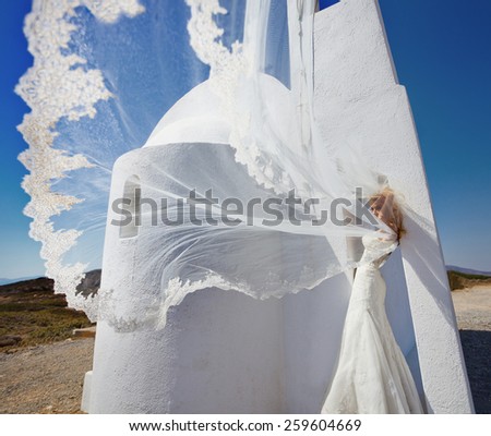 A young beautiful blond bride stands on a fabulous balcony with beautiful wooden roof in a luxury hotel in a white wedding dress unique in Santorini on the background of the sunny sky