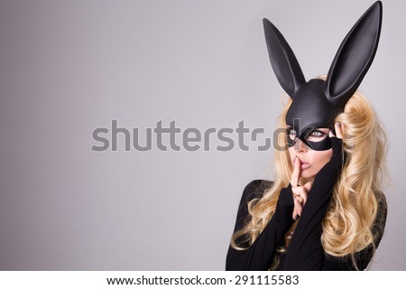 Beautiful blonde-haired young woman in carnival mask ballroom rabbit with long ears sensual sexy in a black dress, standing defiantly on a white background, finger clog the mouth for silence