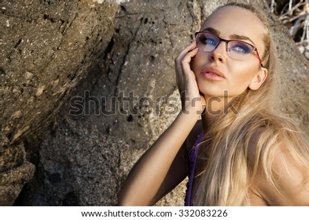 Beautiful blond hair sexy woman young girl model in sunglasses and elegant black swimsuit with crystals on the rocks and wildlife the sea and the island of Santorini