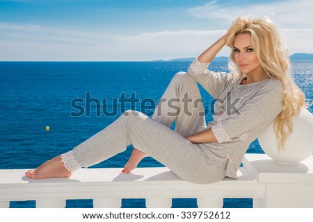Beautiful blond hair model sexy woman standing at the pool of short gray lace pajamas  at the railing on the background blue sea siting balustrade
