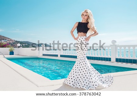 Beautiful blonde young slim woman model girl standing next to an exclusive pool and spa Willas in a long wedding dress carnival ballroom polka-dots in the island of Santorini island spain