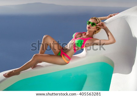 beautiful young blonde woman in a tan color swimsuit lying by the pool on the island of Santorini in Greece, in the background is a beautiful amazing landscape on the mountains and the sea
