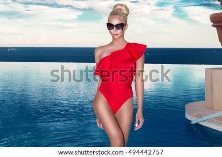 Beautiful blonde woman model with amazing body standing in the pool in an elegant red swimsuit and in the background is amazing view of the sea and sky.