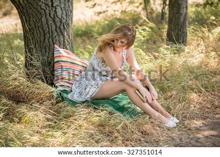 girl sitting in an old oak tree and dreams. In the hands of her book. Wind waves girl\'s hair. Beside her pillow and blanket in the style of boho. The girl light dress. Fain art film.