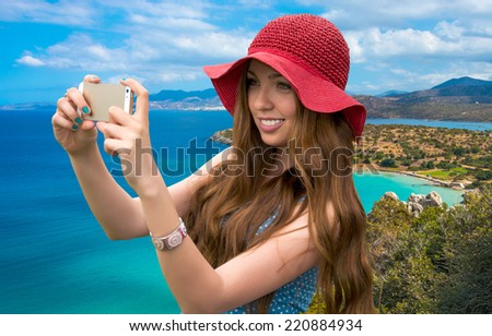 Beautiful woman in a red hat taken picture of herself, selfie. Sea coast in a quiet sunny day. Greece, Crete, Mirabello Bay, Aegean Sea