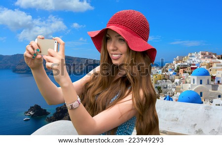 Beautiful woman in a red hat taken picture of herself, selfie in front of traditional architecture of Santorini, sea view.