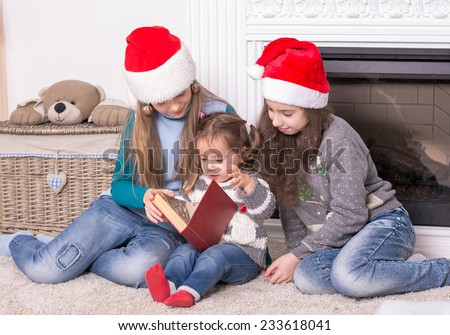 Elder sisters reading a Christmas story his little sister. Girls in Santa\'s hat holding a book near the fireplace.