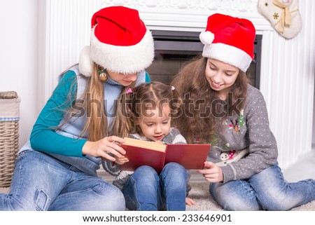 Elder sisters reading a Christmas story his little sister. Girls in Santa\'s hat holding a book near the fireplace.