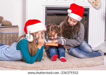 Elder sisters reading a Christmas story his little sister. Girls in Santa's hat holding a book near the fireplace.