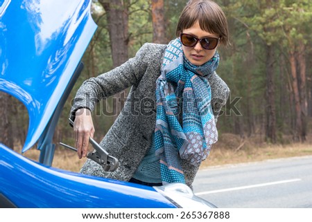 A girl is at a loss with the wrong wrench over the open car hood in the forest.