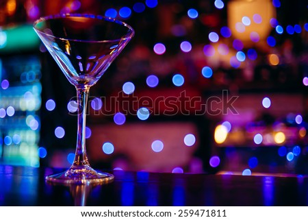 clean empty cocktail glass martini on a dark bar counter with a background of neon lights and bokeh