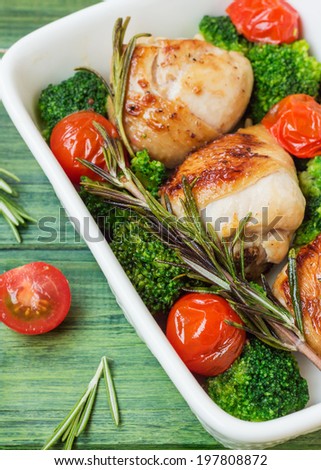 Chicken legs with cherry tomatoes, broccoli, rosemary in a white bowl on the background of the tree.