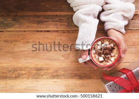 female hand holding cup of hot cocoa or chocolate with marshmallow on wooden table from above