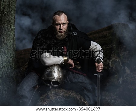 Viking with sword and helmet on a background of smoky forest. Warrior resting. Historic costume.