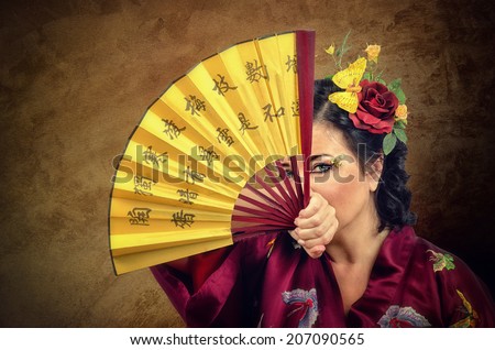 Kimono woman with flowers in hair looking at you through the open Asian fan. Portrait on grunge background